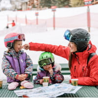 Two kids eating lunch at a table with a ski pro at a ski resort