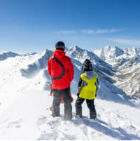 Kid in yellow jacket standing next to a ski pro in a red jacket on a mountain cliff