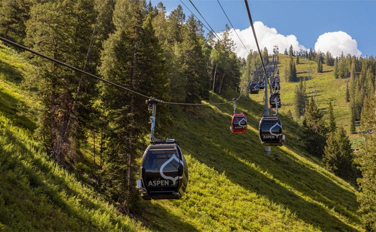 Gondola cars travel up Aspen Mountain on a sunny summer afternoon day, the sun lasts long shadows through the pine trees, and the gondola cars travel over green meadows