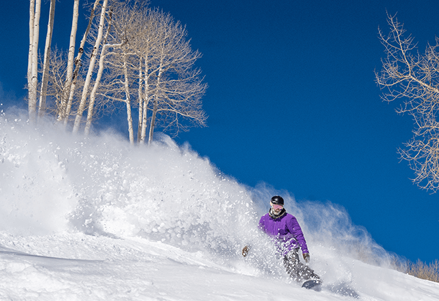 Man wearing purple jacket snowboards through deep, very white powder. The blue sky is bright behind him at Aspen Snowmass