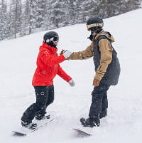 Snowboarder taking a lesson with a ski pro in a red jacket on a ski mountain