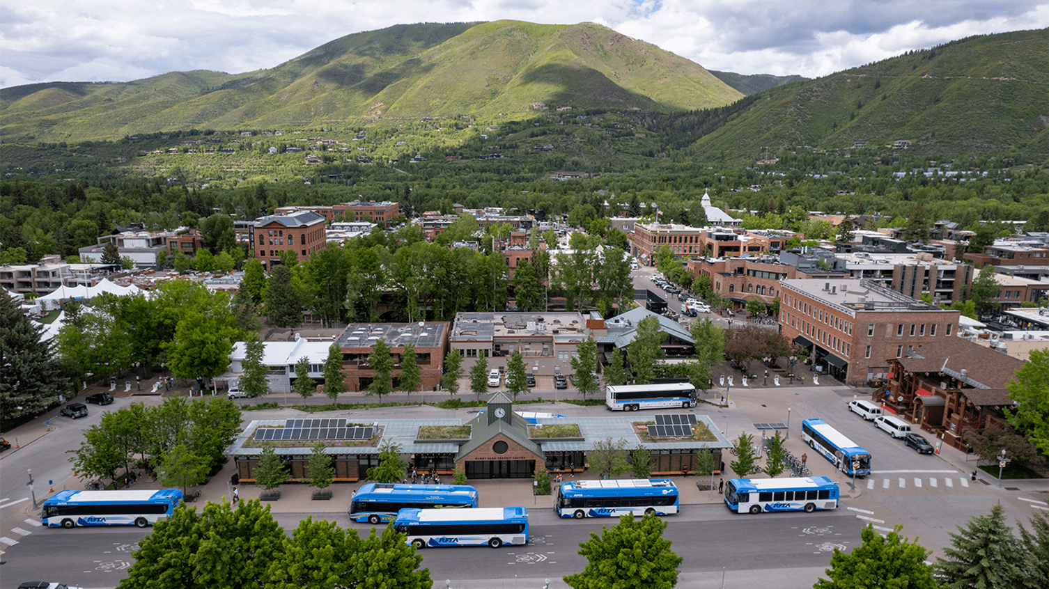 Aerial shot of many RFTA buses parked and pulling out of the Ruby Stop in the Heart of Aspen,  town surrounds the bus stop, and green mountains can be seen in the background