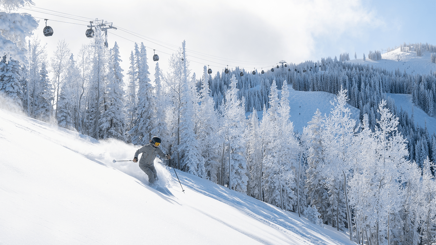 Skier in deep powder on Aspen Mountain, under the gondola. Blue skies above snowy trees and powder.