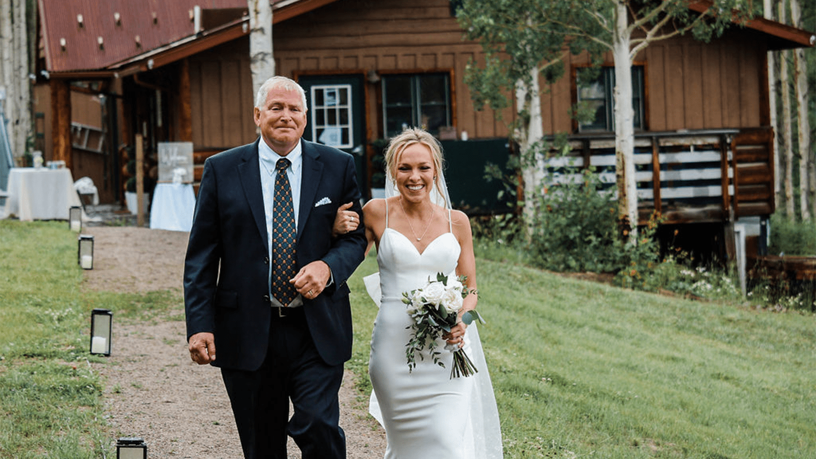 Bride walking down the Isle with her father, green grass and small mountain cabin, wit aspen trees, in background